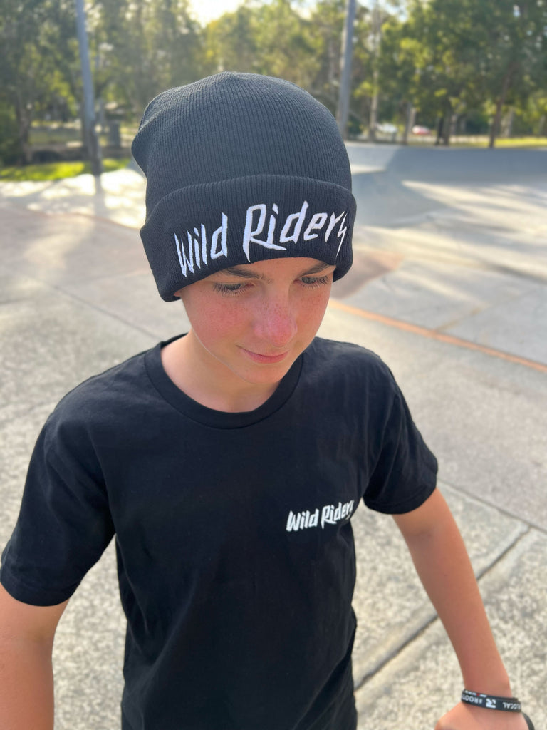 Archie scooter rider wearing a WIld Riders black tshirt and black Wild Riders beanie hat, standing at an outdoor skatepark