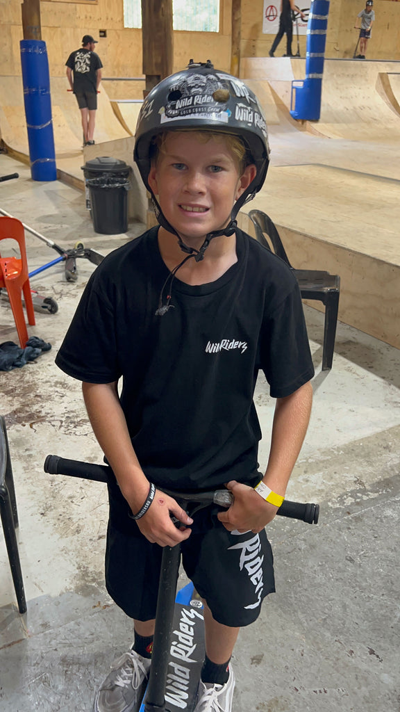 EJ scooter rider wearing wild riders tshirt, shorts, wristband and has a wild riders griptape on his scooter. He is at Ramp Attak indoor skatepark with helmet on and Wild Riders stickers on his helmet.